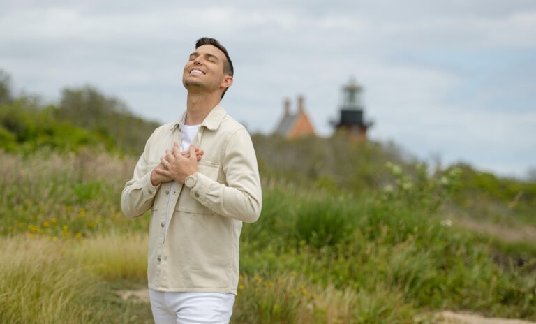 Matt Fraser, dressed in a light-colored outfit, is standing outdoors with his eyes closed and hands clasped to his chest. A lighthouse stands visible in the background amidst the greenery.