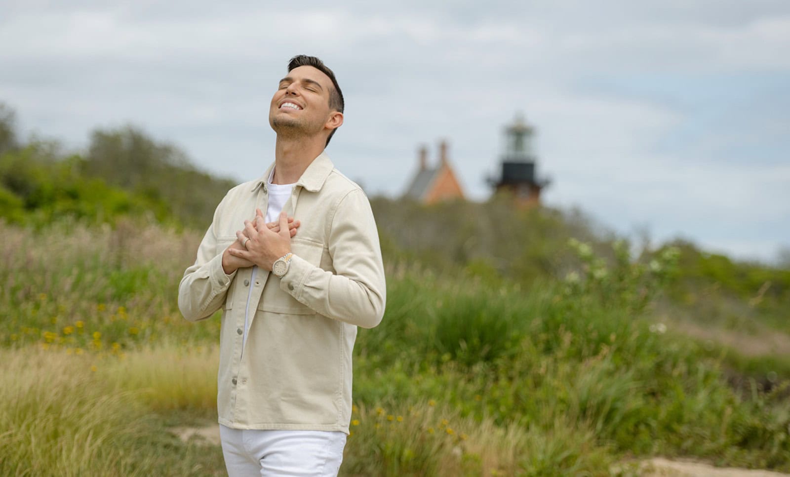 Matt Fraser, dressed in a light-colored outfit, is standing outdoors with his eyes closed and hands clasped to his chest. A lighthouse stands visible in the background amidst the greenery.