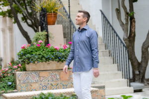 Matt Fraser in a blue button-up shirt and white pants walks outdoors near tiled steps, potted plants, and flowers.