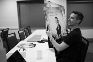 A person named Matt Fraser is sitting at a table holding large posters in a conference room, with pens and a cup nearby.