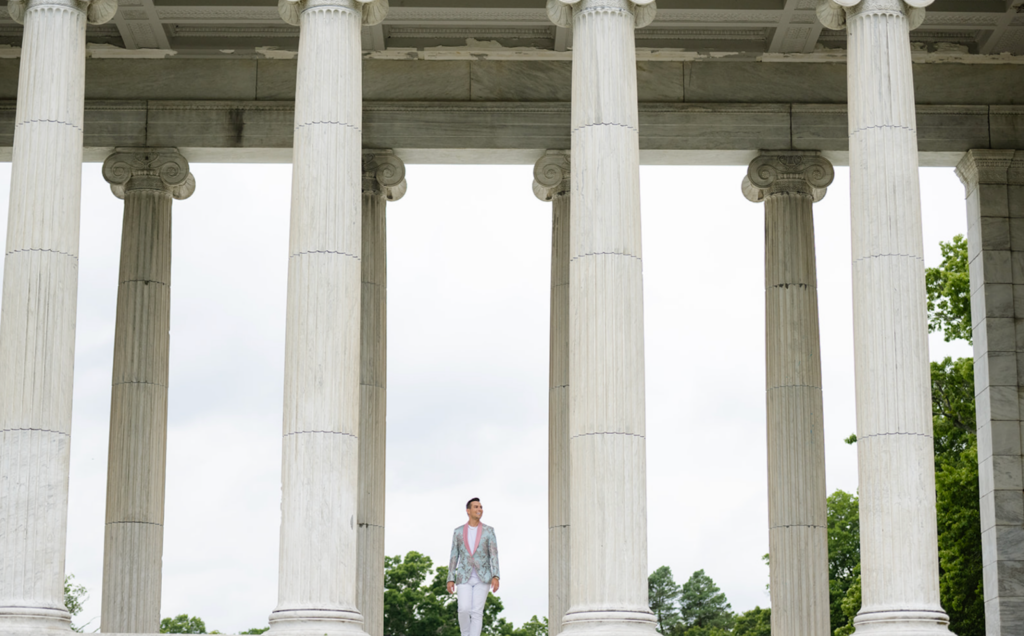 Matt Fraser, in formal attire, stands between tall, classical columns with a cloudy sky and greenery in the background.