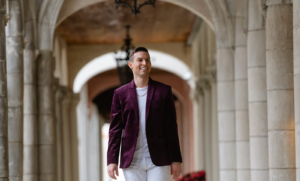 Matt Fraser, dressed in a purple blazer and white pants, walks through a stone archway with a neutral expression.