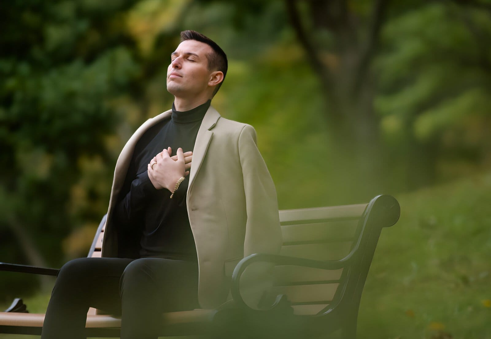 Matt Fraser in a beige coat sits on a park bench with eyes closed, one hand on their chest. Green foliage is visible in the background.