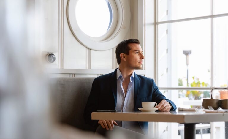 Matt Fraser, dressed in a suit, sits at a table with a cup of coffee, looking out a large window in a bright, modern cafe.