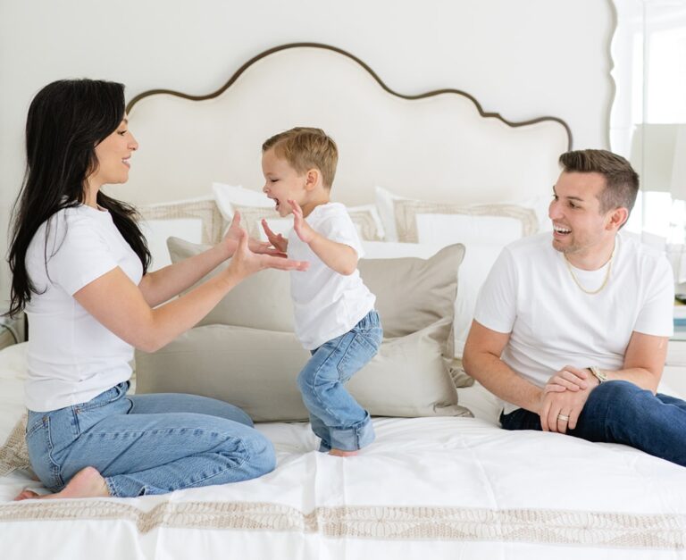 A woman and Matt Fraser watch a child standing on a bed. All three are wearing white shirts and jeans, and appear to be enjoying a playful moment together.