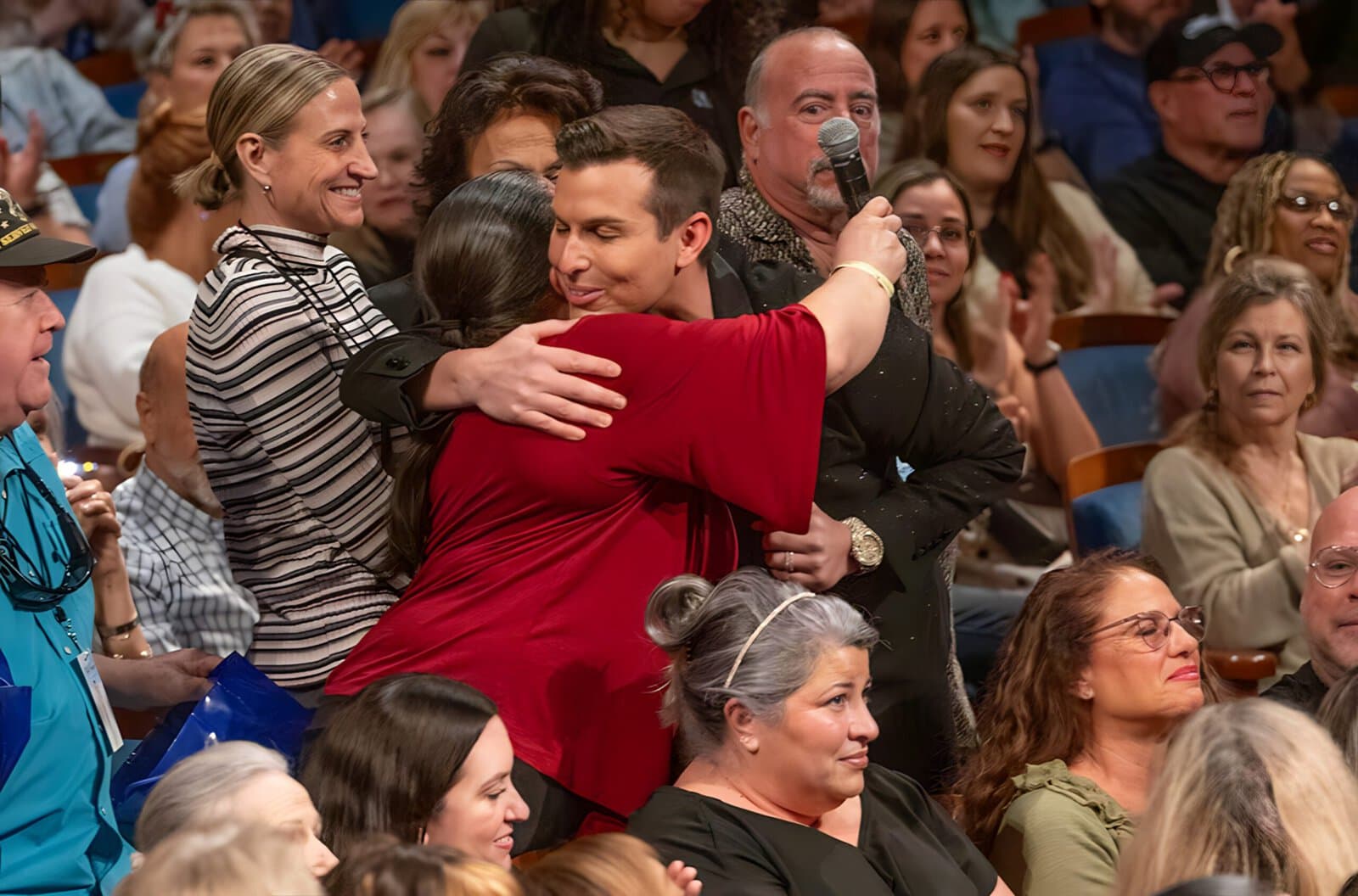 A woman in red hugs Matt Fraser, who is holding a microphone, in a crowded auditorium. Other seated audience members watch the interaction.