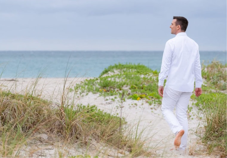 Matt Fraser, dressed in white clothing, walks barefoot on a sandy beach with greenery, looking towards the ocean under a cloudy sky.