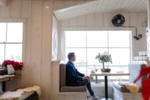 Matt Fraser is sitting in a booth by a large window in a bright café, with plants on the table and wooden walls surrounding the area.