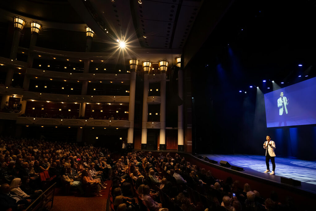 Matt Fraser stands on stage, speaking to a large audience in a darkened theater. A screen to the right projects his image.