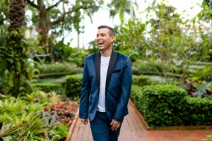 Matt Fraser, dressed in a blue suit and white shirt, is walking and smiling on a brick path in a lush garden.