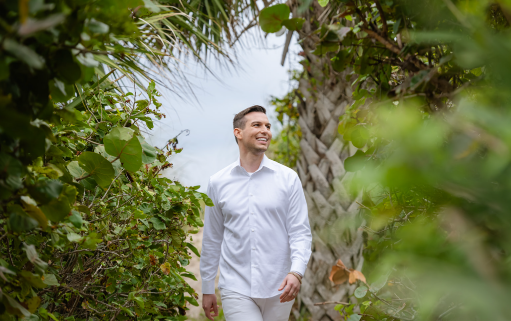 A man named Matt Fraser, wearing a white shirt, walks outside, surrounded by lush greenery and a palm tree.