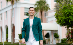 Matt Fraser, wearing a teal blazer and white pants, smiles while walking outside a pastel-colored building with palm trees.