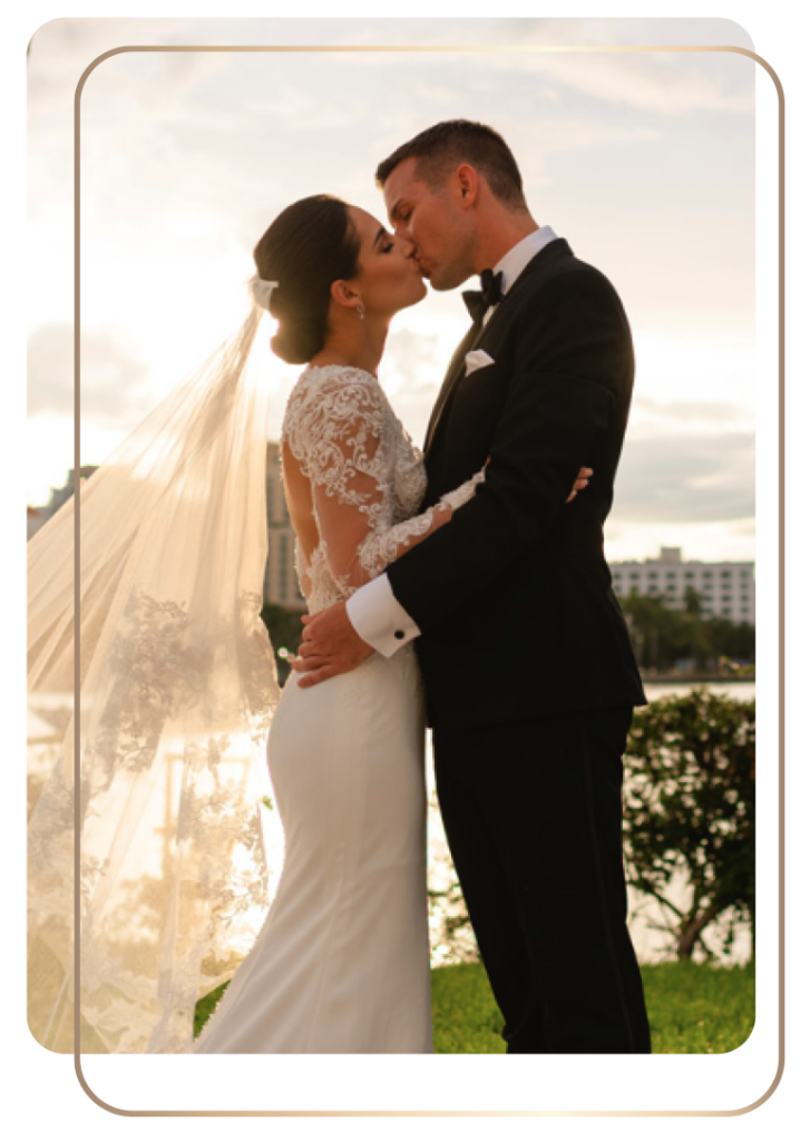 Matt & Alexa Fraser share a kiss outdoors during sunset. Alexa is in a white dress with a long veil; Matt Fraser is in a black tuxedo. They are embracing near a water body with buildings in the background.