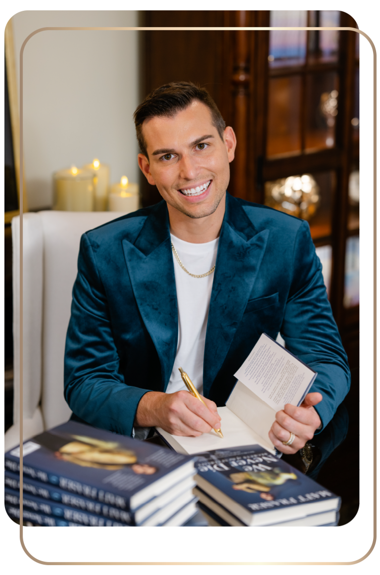 Matt Fraser in a blue jacket sits at a table, smiling and signing a book with a pen. Several copies of the book are stacked on the table.