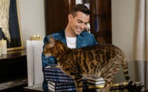 Matt Fraser, in a blue jacket, sits at a table with stacks of books, smiling. A cat walks across the table in front of him.