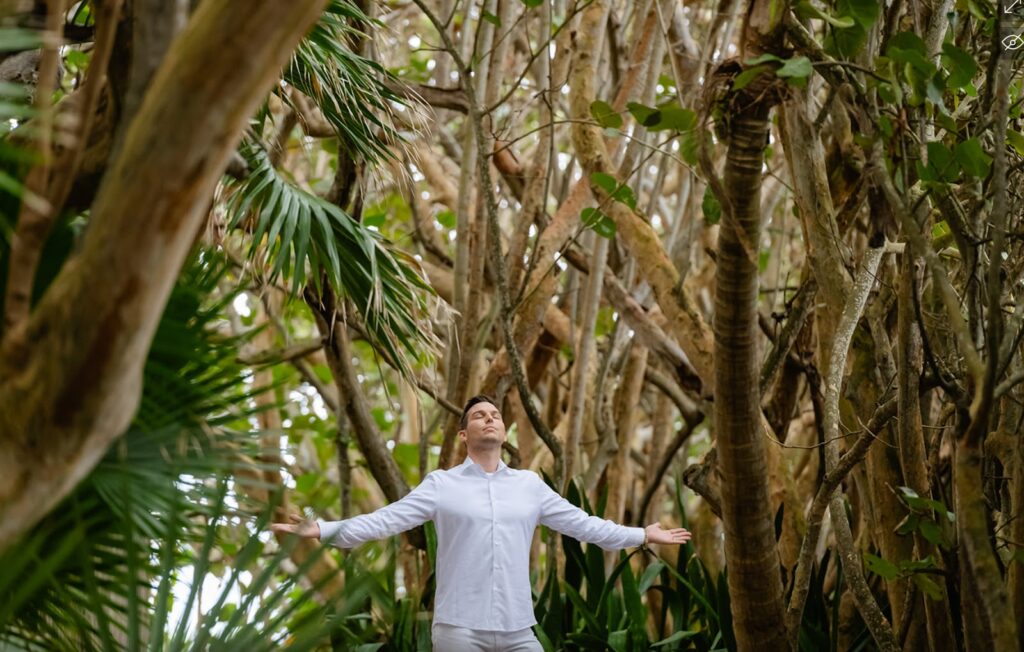 A person in a white outfit, possibly Matt Fraser, stands with arms outstretched, surrounded by tall, dense trees and greenery.