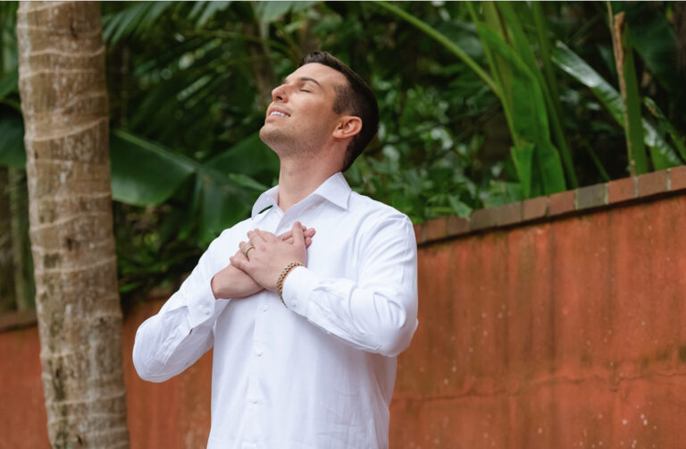 Matt Fraser, in a white shirt, stands outdoors with closed eyes and hands on chest, appearing content. A brick wall and lush greenery are in the background.