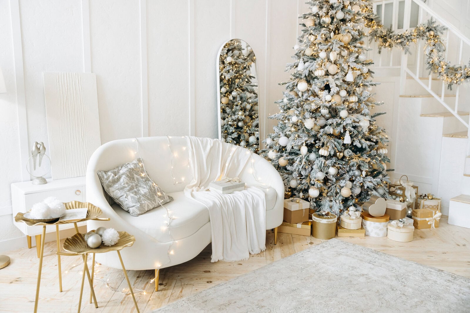 Elegant living room with a white curved sofa, a decorated Christmas tree, and gold-accented decor. Presents are arranged under the tree, and soft lighting adds warmth to the scene.