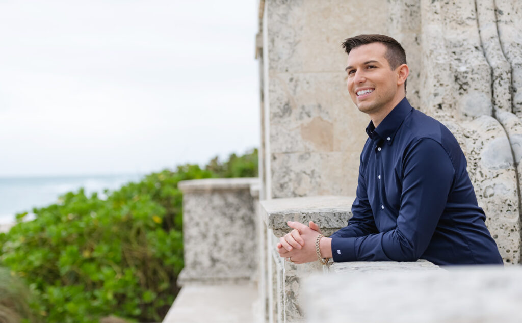 Matt Fraser, wearing a navy shirt, leans against a stone ledge, smiling, with greenery and the ocean in the background.