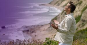 Matt Fraser stands outdoors with his eyes closed and arms slightly outstretched at a cliffside beach with waves and hills in the background.