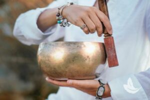 Matt Fraser, wearing bracelets, holds a singing bowl in one hand and uses a mallet to produce sound with the other hand.