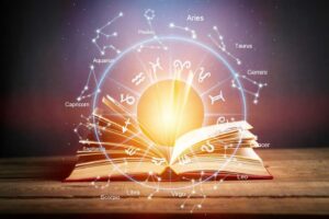 An open book on a wooden table is illuminated by a bright light. Surrounding it are zodiac signs and constellations in a circular pattern, labelled with their respective names.