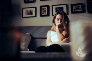 A woman sits on a couch in a dimly lit room with framed photos on the wall behind her. She appears deep in thought, with one hand covering her mouth and the other arm wrapped around her knee, reflecting on memories that include Matt Fraser.