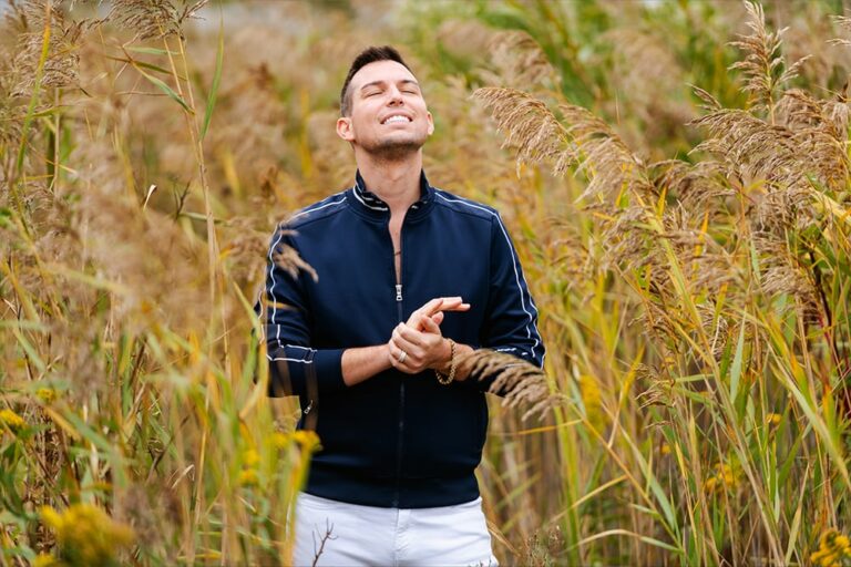 Matt Fraser stands in a field of tall grass, wearing a navy jacket and white shorts, looking upwards with eyes closed and hands clasped.