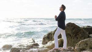 Matt Fraser stands on the rocky shore, facing the ocean with hands in prayer position. He is wearing a dark shirt and white pants, with waves in the background.